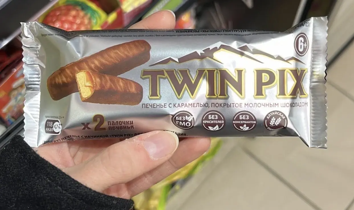 A photo of a russian twix-knockoff candy bar. The packaging is titled "Twin Pix", it depicts a pair of twix-like caramel cookie candy with silver mountain peaks in the background. The person taking the photo is holding the candy bar in their hand. Grocery store shelves are visible in the background.