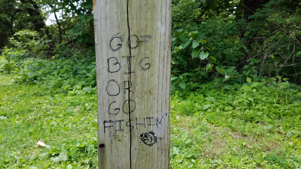 Closeup of a cracked, grayish brown wooded post surround by grass with some brush in the background. Written on the post in permanent marker are the words: &quot;Go big or go fishin&quot;. There is also a cartoony drawing of a fish with big cheeks, big eyes, and plewds.