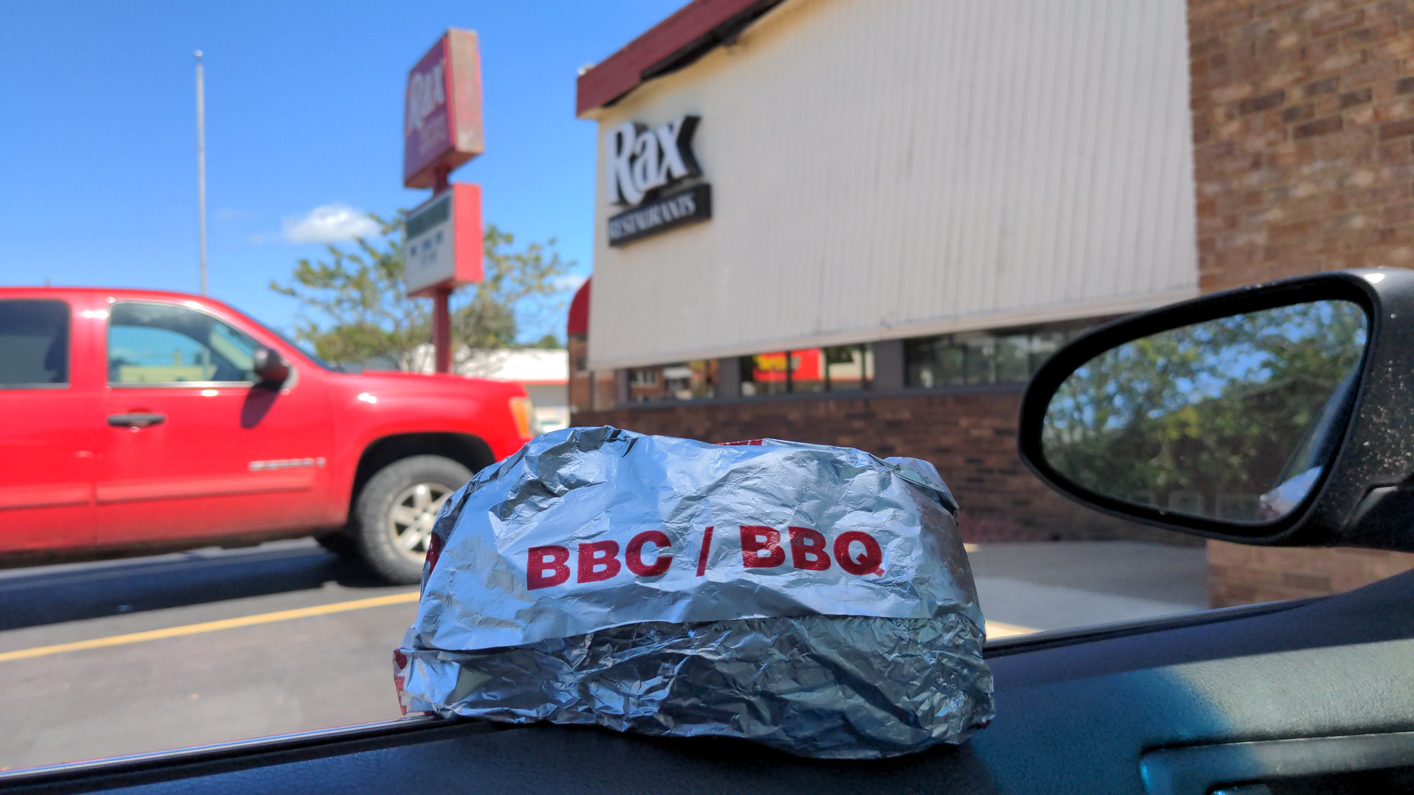 A sandwich wrapped up in silver foil with the red letters BBC and BBQ on the outside. The sandwich sits on the ledge of a rolled down car window. Outside the window is a fast food restaurant named Rax.